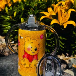 K&ouml;nnte beinhalten: Ein gelber, glitzernder Schnabelbecher mit einer Grafik von Winnie Puuh im roten T-Shirt, begleitet von kleinen Bienen- und Blumenillustrationen. Der Becher verf&uuml;gt &uuml;ber zwei transparente Kunststoffgriffe, einen Schnabeldeckel und einen Zusatzdeckel.