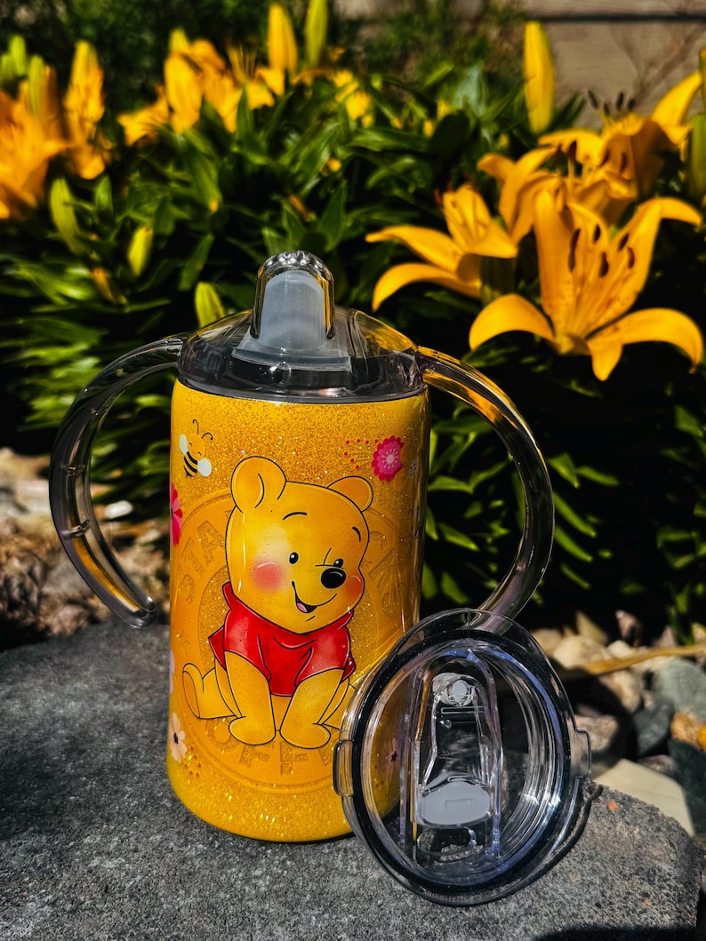 K&ouml;nnte beinhalten: Ein gelber, glitzernder Schnabelbecher mit einer Grafik von Winnie Puuh im roten T-Shirt, begleitet von kleinen Bienen- und Blumenillustrationen. Der Becher verf&uuml;gt &uuml;ber zwei transparente Kunststoffgriffe, einen Schnabeldeckel und einen Zusatzdeckel.