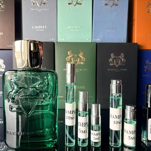 May include: A collection of Parfums de Marly fragrances, including a large green bottle and several sample vials. The bottles are arranged in front of various boxes with gold lettering and crests. The samples are labeled with sizes.