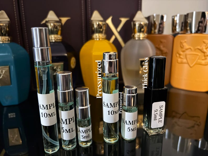 Pu&ograve; includere: Un assortimento di campioni di profumo in flaconi di vetro trasparente con tappi argentati. I campioni sono etichettati con dimensioni come 10 ml, 5 ml e 3 ml. Diverse bottiglie di profumo di dimensioni standard in vari colori sono visibili sullo sfondo.
