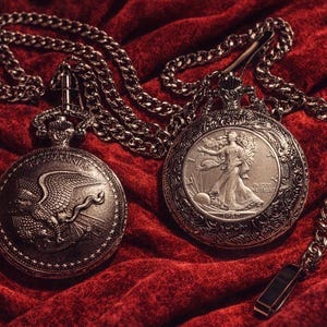 May include: Two antique silver pocket watches with chains rest on a red velvet surface. One watch features an eagle design, the other a woman. The chains and watch casings have a tarnished, vintage appearance.