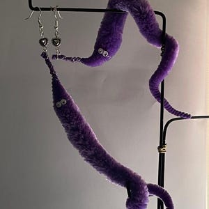 May include: A pair of quirky purple worm earrings with silver heart charms. The earrings are displayed on a black metal stand, with a matching purple worm. The worms have googly eyes and a fuzzy texture.