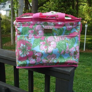 K&ouml;nnte beinhalten: Rechteckige K&uuml;hltasche mit einem floralen Muster in Rosa, Blau und Gr&uuml;n. Die Tasche hat rosa Einfassungen, Griffe und eine Vordertasche. Sie steht auf einem Holzgel&auml;nder, mit gr&uuml;nem Hintergrund.