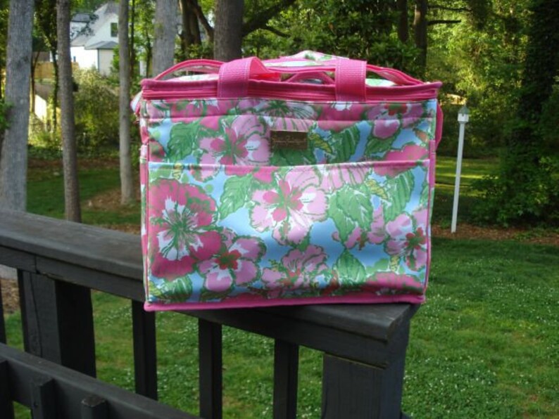 K&ouml;nnte beinhalten: Rechteckige K&uuml;hltasche mit einem floralen Muster in Rosa, Blau und Gr&uuml;n. Die Tasche hat rosa Einfassungen, Griffe und eine Vordertasche. Sie steht auf einem Holzgel&auml;nder, mit gr&uuml;nem Hintergrund.