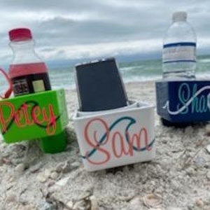 May include: Three small, personalized containers on a sandy beach. One green container with "Petey" in red script holds a bottle of juice and glasses. A white container with "Sam" in teal script holds a phone. A blue container with "Sharon" in white script holds a water bottle.