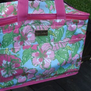 K&ouml;nnte beinhalten: Eine Tragetasche mit floralem Muster, mit einem rosa Rei&szlig;verschluss und Griffen. Die Tasche zeigt ein lebendiges Design aus rosa und gr&uuml;nen Blumen auf hellblauem Hintergrund. Ein kleines, goldfarbenes rechteckiges Etikett ist sichtbar.