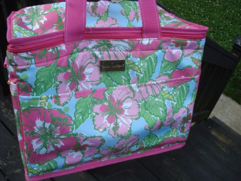 K&ouml;nnte beinhalten: Eine Tragetasche mit floralem Muster, mit einem rosa Rei&szlig;verschluss und Griffen. Die Tasche zeigt ein lebendiges Design aus rosa und gr&uuml;nen Blumen auf hellblauem Hintergrund. Ein kleines, goldfarbenes rechteckiges Etikett ist sichtbar.