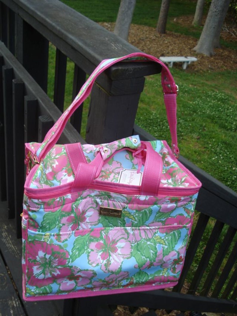 K&ouml;nnte beinhalten: Eine Tragetasche mit rosa-blauem Blumenmuster und passendem Schulterriemen. Die Tasche hat einen Rei&szlig;verschluss oben, eine Vordertasche und ein goldfarbenes Logo. Das Blumenmuster umfasst rosa Bl&uuml;ten und gr&uuml;ne Bl&auml;tter auf hellblauem Hintergrund.