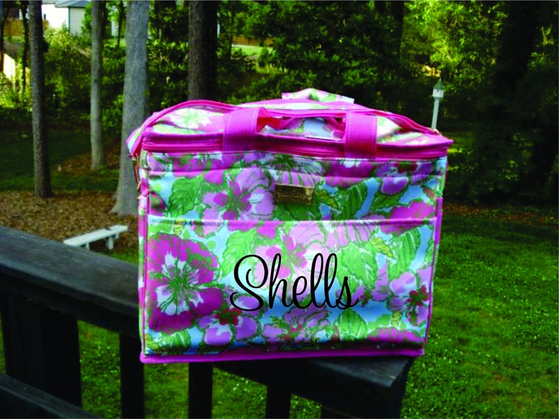 K&ouml;nnte beinhalten: Eine Tragetasche mit floralem Muster, rosa Besatz und Griffen. Die Tasche hat ein Design aus rosa und lila Blumen mit gr&uuml;nen Bl&auml;ttern auf hellblauem Hintergrund. Das Wort "Shells" ist auf der Vorderseite geschrieben.