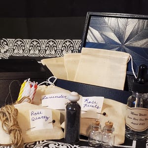 May include: An open black box with a geometric design, filled with small canvas bags, bottles, and candles. The bags are labeled with handwritten tags: "Rose Quartz", "Lavender", and "Rose Petals". A bottle of "Moon Water" is also visible.