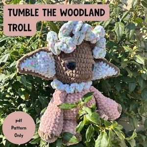 Peut inclure: Une poupée troll au crochet faite à la main avec le texte "Tumble the Woodland Troll". La poupée a une tête marron, un corps rose et des accents blancs et roses. La poupée est entourée de verdure. Le texte "pdf Pattern Only" est également visible.