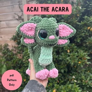 Puede incluir: Un peluche de ganchillo verde con detalles rosas, que representa una criatura con ojos grandes y una bufanda verde claro. El texto "ACAI THE ACARA" se muestra encima del juguete, con "pdf Pattern Only" visible debajo. El juguete se sostiene en una mano.
