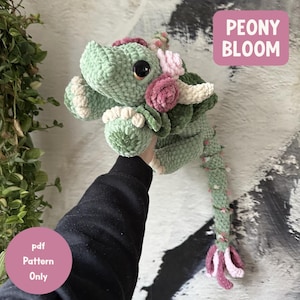May include: A crocheted dragon in shades of green, pink, and cream. The dragon has large black eyes and is adorned with pink and cream-colored flowers. The text "PEONY BLOOM" is in a pink box. A pink circle with the text "pdf Pattern Only" is also visible.