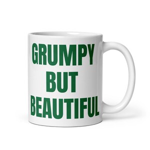 Könnte beinhalten: Weiße Keramiktasse mit den Worten "GRUMPY BUT BEAUTIFUL" in fetten grünen Buchstaben. Die Tasse hat einen gebogenen Griff und eine glänzende Oberfläche. Ein schlichtes, aber auffälliges Design für eine Kaffee- oder Teetasse.