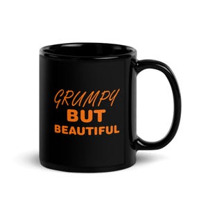 Könnte beinhalten: Schwarze Keramik-Tasse mit gebogenem Henkel. Die Tasse zeigt den orangefarbenen Text "GRUMPY BUT BEAUTIFUL" in einer verspielten Schriftart. Die Tasse ist für heiße oder kalte Getränke konzipiert.