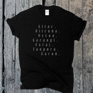 May include: Black t-shirt with a list of musical instruments printed in a light gray color. The instruments listed are Sitar, Dilruba, Veena, Sarangi, Esraj, Tanpura, and Sarod. The shirt is displayed on a wooden surface.