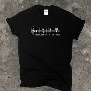 May include: Black t-shirt featuring a white musical staff design with notes and the text "THESE ARE DIFFICULT TIMES". The shirt has a crew neck and is made of a soft material.