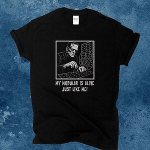 May include: Black t-shirt featuring a black and white graphic of Frankenstein's monster working on a modular synthesizer. The text "MY MODULAR IS ALIVE JUST LIKE ME!" is printed below the image.