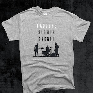 May include: A grey t-shirt featuring the words "SADCORE SLOWER SADDER" in white. Beneath the text is a black silhouette of a band performing. The t-shirt is made of a soft fabric.