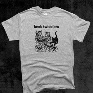 May include: A grey t-shirt with the words "knob twiddlers" above a black and white illustration of three cats interacting with electronic music equipment. One cat is black.