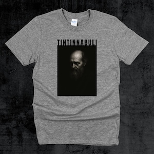 May include: A grey t-shirt with a black and white portrait of a bearded man, and the word "TINTINNABULI" in a black, bold font above the portrait. The shirt is made of a soft material.