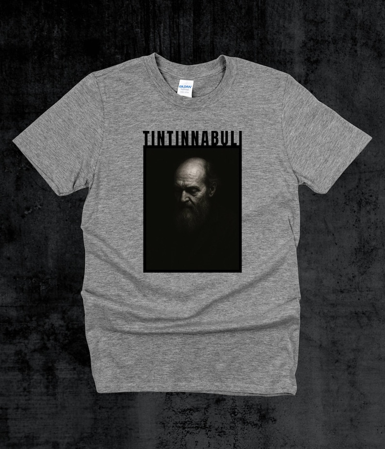 May include: A grey t-shirt with a black and white portrait of a bearded man, and the word "TINTINNABULI" in a black, bold font above the portrait. The shirt is made of a soft material.