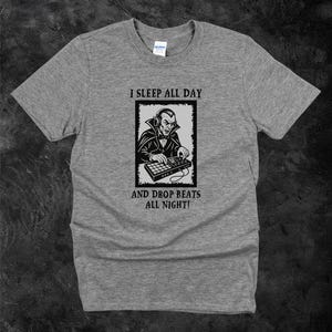 May include: A heather gray t-shirt featuring a black and white graphic of a vampire DJ with headphones and a mixing board. The text reads "I SLEEP ALL DAY AND DROP BEATS ALL NIGHT!"
