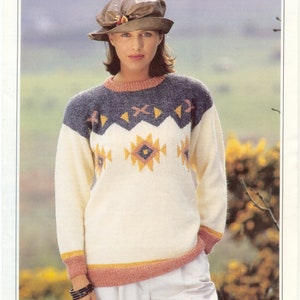 May include: A woman models a cream-coloured sweater with a geometric pattern in yellow, orange, and blue. The sweater features a navy blue yoke and pink and yellow trim. The image also shows a brown hat and white trousers.