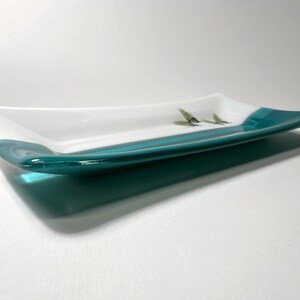 May include: A rectangular glass serving dish with a white interior and a teal blue rim. The dish is slightly curved and has a glossy finish.