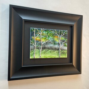 Puede incluir: Una obra de arte enmarcada que representa una escena de bosque con tres &aacute;rboles en primer plano. Los &aacute;rboles est&aacute;n silueteados contra un fondo verde y azul con toques de amarillo y naranja. La obra de arte est&aacute; enmarcada en un marco negro.