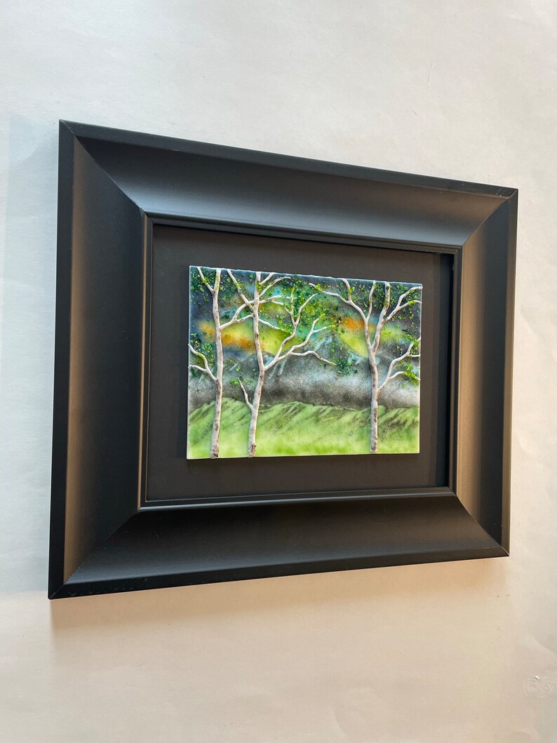 Puede incluir: Una obra de arte enmarcada que representa una escena de bosque con tres &aacute;rboles en primer plano. Los &aacute;rboles est&aacute;n silueteados contra un fondo verde y azul con toques de amarillo y naranja. La obra de arte est&aacute; enmarcada en un marco negro.