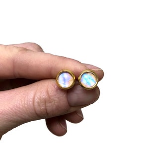 Puede incluir: Un par de pendientes de botón dorados. Cada pendiente presenta un diseño redondo, iridiscente y multicolor con tonos de azul, morado y amarillo. Los pendientes se sostienen en una mano sobre un fondo blanco.