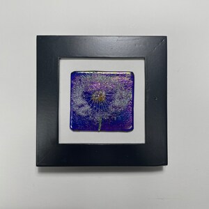 Op de afbeelding: Een ingelijst glazen kunstwerk met een witte paardenbloem met een gouden hart op een paarse, iriserende achtergrond.