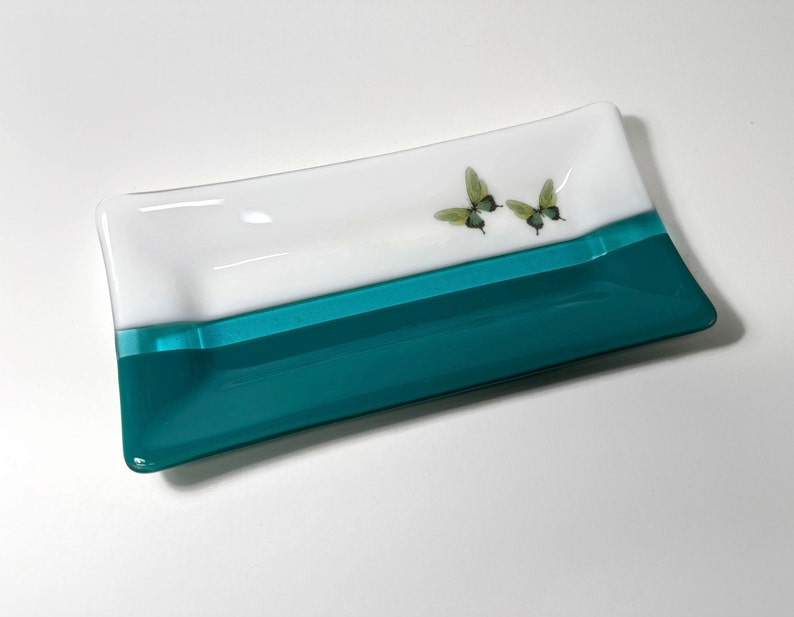 May include: A rectangular glass dish with a white top and a teal bottom. Two green butterflies are painted on the white surface.