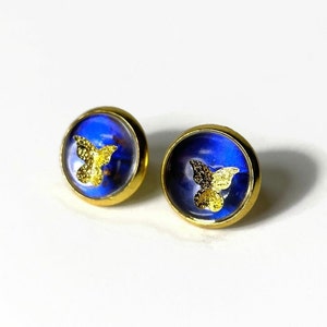 Puede incluir: Un par de pendientes de botón dorados con fondo azul y diseño de mariposa dorada.