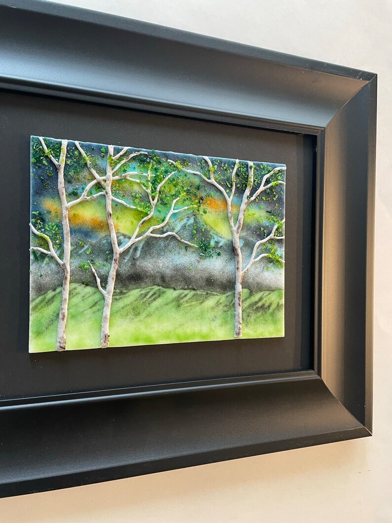 Puede incluir: Obra de arte enmarcada que presenta un mosaico de vidrio verde, azul y blanco que representa un paisaje con dos abedules y una colina verde.