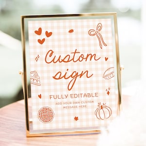 May include: A rectangular sign in a gold metal frame featuring a light orange and white gingham background. The text reads Custom sign, FULLY EDITABLE, and ADD YOUR OWN CUSTOM MESSAGE HERE, surrounded by orange line-art icons of hearts, a bow, and a pumpkin.