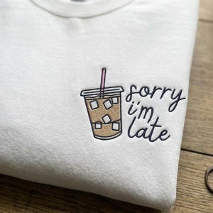 以下が含まれることがあります： ピンクのストローと氷が入ったプラスチックカップのアイスコーヒーの刺繍が施された白いクルーネックのスウェットシャツ。カップの横には、濃い青色の筆記体で sorry i'm late という刺繍文字がデザインされています。