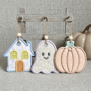 May include: Three hanging ornaments featuring a house with yellow windows, a ghost with a spiderweb pattern, and a peach-colored pumpkin. Each ornament is crafted with embroidered details and hangs from a natural twine loop with a wooden bead.