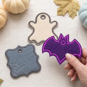 May include: Three felt gift tags in different shapes and colors. One is a cream-colored ghost shape, one is a slate blue square shape with a stitched spiderweb design, and one is a purple bat shape with a dark purple embroidered border.