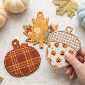 Puede incluir: Tres adornos de tela con forma de hoja de arce, calabaza y bellota. La hoja de arce es de color amarillo mostaza con un estampado floral. La calabaza es naranja con un diseño de cuadros. La bellota tiene una parte superior de cuadros vichy beige.