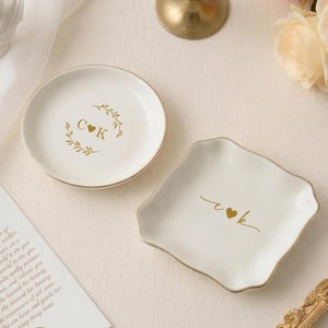 May include: Two white ceramic ring dishes with gold trim. One is round with a gold leaf design and the initials "C K". The other is square with the initials "c♥k". Wedding theme.