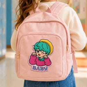 Peut inclure: Un sac à dos rose clair avec un écusson brodé central représentant un personnage de dessin animé aux cheveux bleu sarcelle, portant un béret jaune et un pull rose. Le personnage fait un clin d'œil. Le mot BABY est brodé en bleu en dessous.