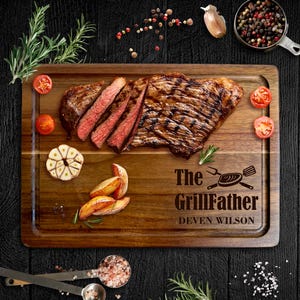 Può includere: Un tagliere in legno con bordo intagliato, con una bistecca a fette, patate arrosto, pomodori e aglio. La tavola è incisa con "The GrillFather" e un nome. Guarnito con rosmarino e grani di pepe.