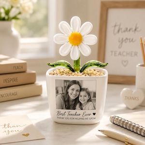 May include: A white ceramic planter featuring a black and white photograph of a woman and a child. The planter has the text "Best Teacher Ever" and "Thank you for helping me grow" printed on the front. A glass daisy with a yellow center sits inside.