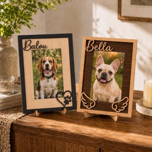 May include: Two rectangular wooden picture frames standing on small supports. The left frame has a black border, the name Balou, and a heart with a paw print. The right frame has a dark brown border, the name Bella, and two angel wings.