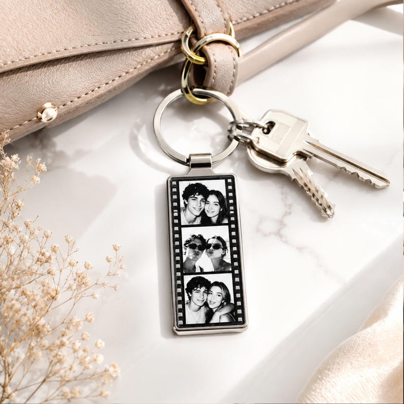 May include: A rectangular silver-toned metal keychain featuring a vertical film strip design. The center contains three stacked black and white photographs of a couple. The keychain is attached to a circular split ring with two silver metal keys.