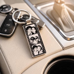 May include: A rectangular metal keychain featuring a vertical film strip design with three black and white photographs of two people. The keychain is attached to a silver metal ring and is positioned next to a black car key fob.