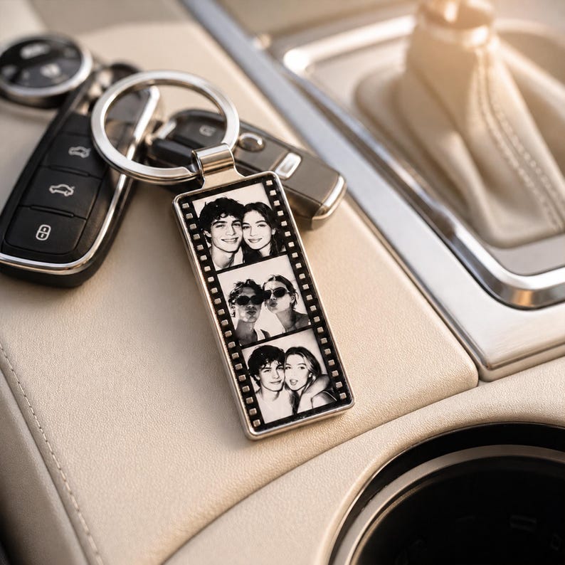 May include: A rectangular metal keychain featuring a vertical film strip design with three black and white photographs of two people. The keychain is attached to a silver metal ring and is positioned next to a black car key fob.