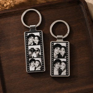 Pode incluir: Dois porta-chaves retangulares de metal com um design de tira de filme a preto e branco. Cada porta-chaves apresenta três retratos sobrepostos de um casal. Os porta-chaves estão presos a argolas de metal prateado para as chaves.